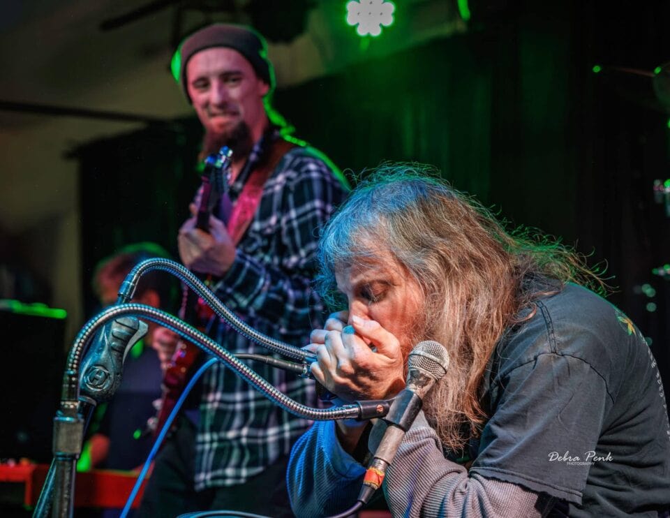 Johnny Wheels playing a harmonica solo with guitar player Brandon Logan approvingly watching.